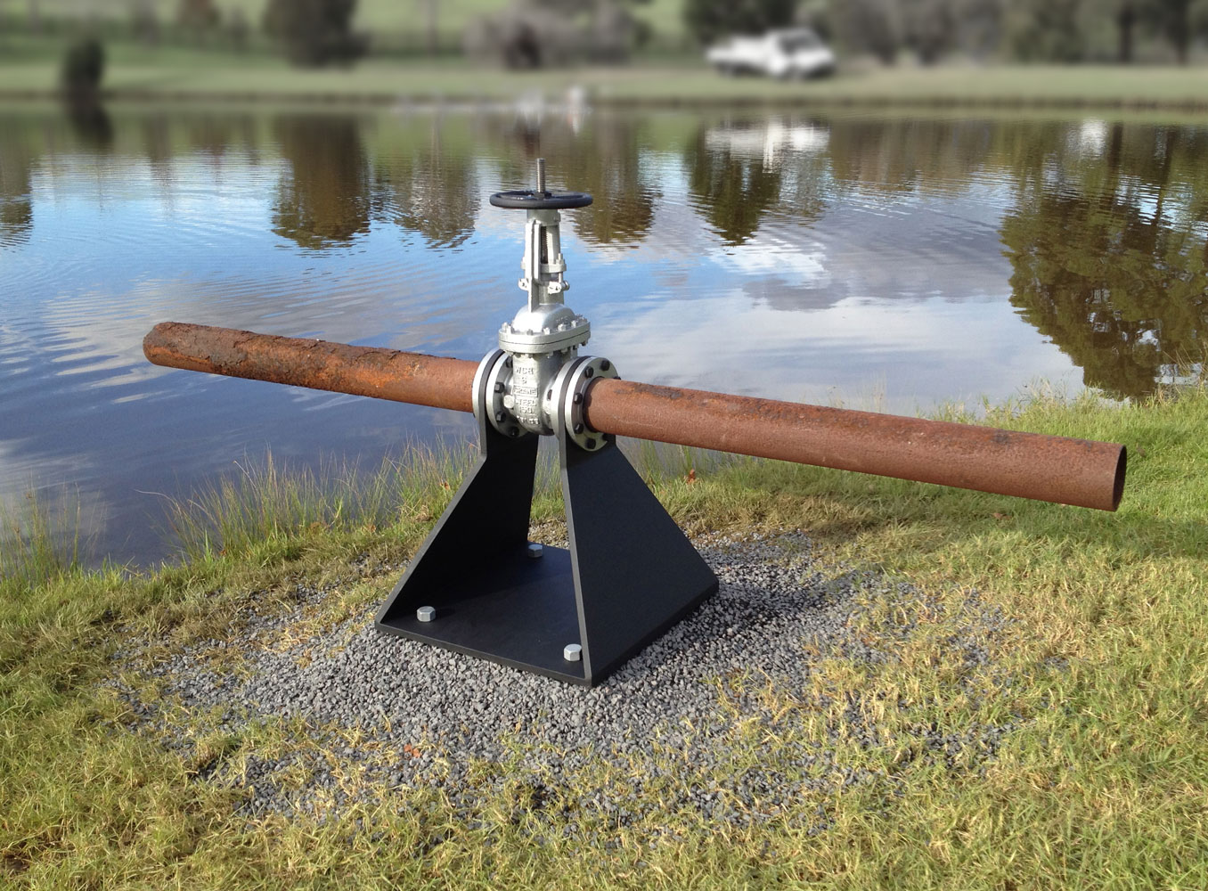 A large steel sculpture, consisting of a four-sided pyramid base, with two two sides empty, supporting a large valve, throgh which a corroded steel pipe projects.